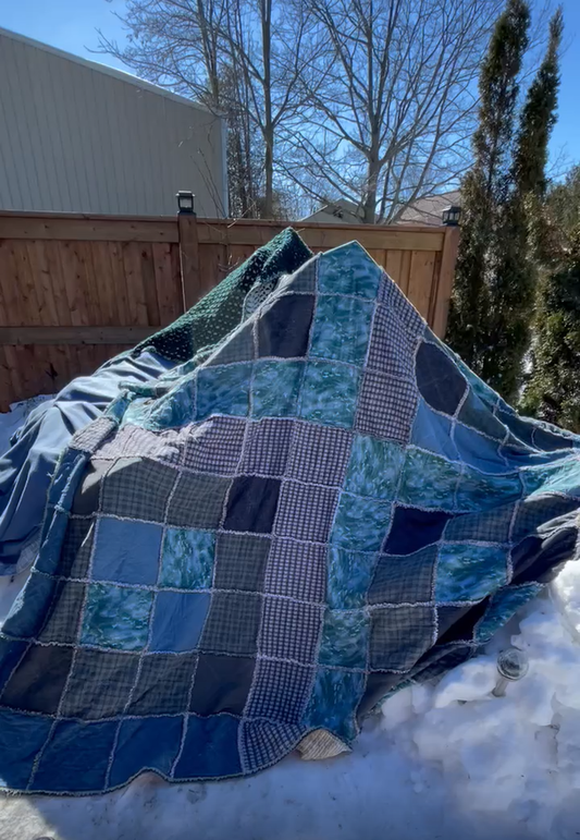 A blanket fort in a snowy rural community. The blue and white patchwork quilt drapes over wood beams.